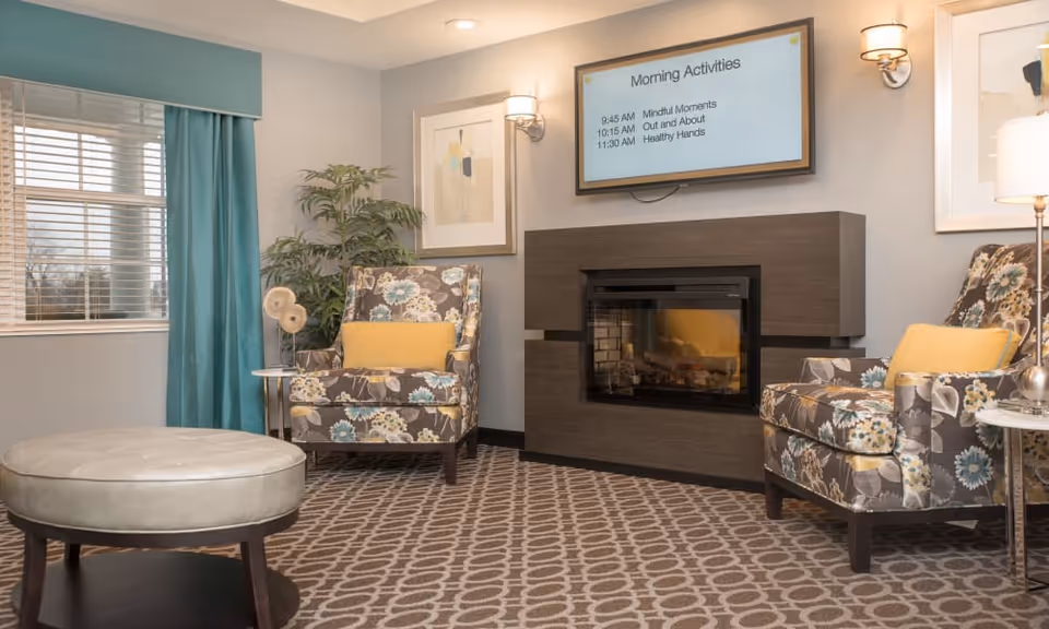 A cozy living room area with two floral-patterned armchairs each with a yellow cushion, a round ottoman, a modern fireplace, a wall-mounted screen displaying morning activities, framed artwork, a potted plant, and teal curtains covering a window.