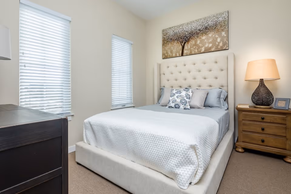 A neatly made bed with a tufted headboard in a bright bedroom featuring a nightstand with lamp, dresser, and two windows with blinds.