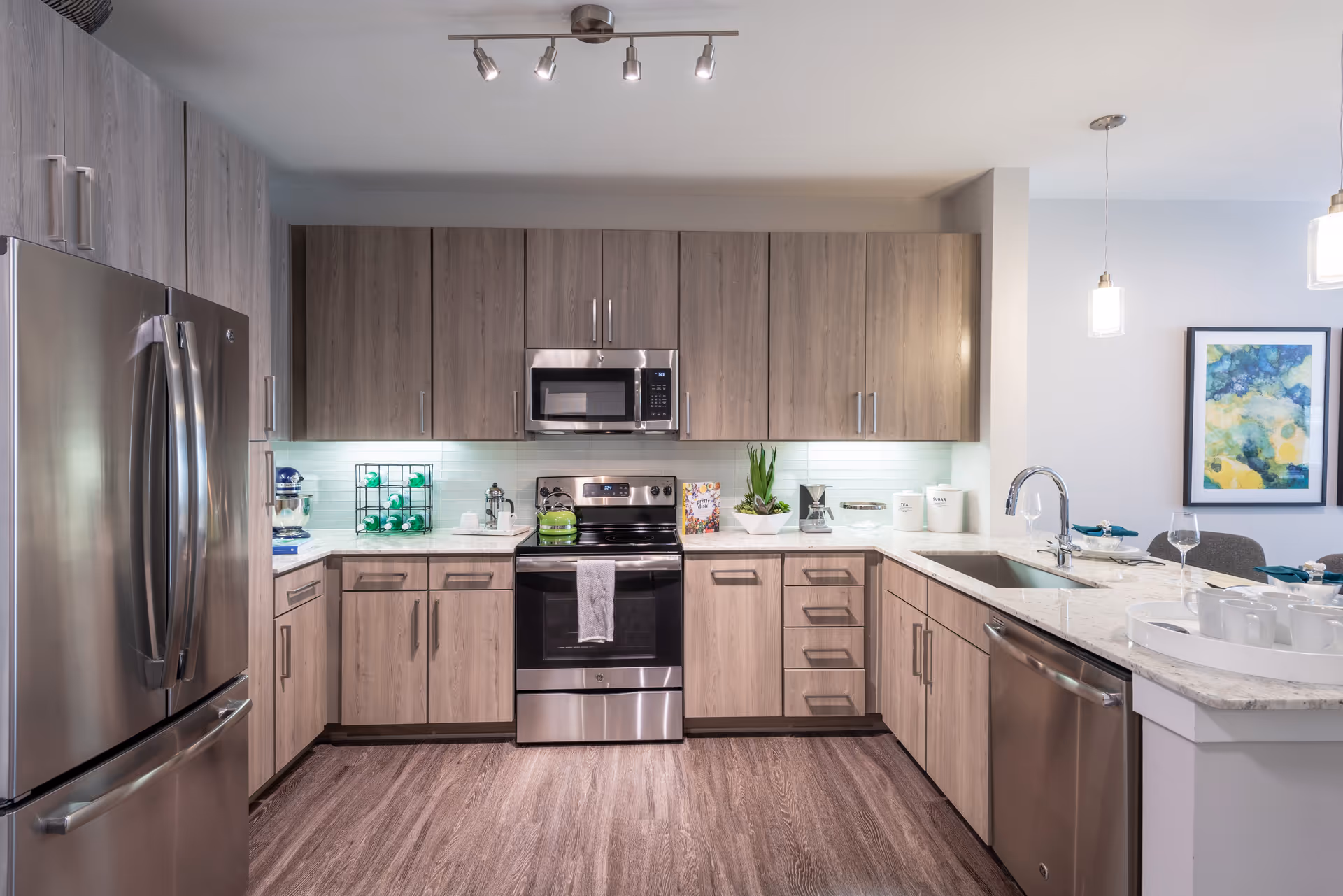 Modern kitchen with stainless steel appliances including a refrigerator, oven, microwave, and dishwasher. Light wood cabinets and drawers surround the appliances, with a white marble countertop and a sink on the right. The kitchen is well-lit with ceiling track lights and two pendant lights hanging over the counter. A colorful abstract painting is visible on the wall in the background.