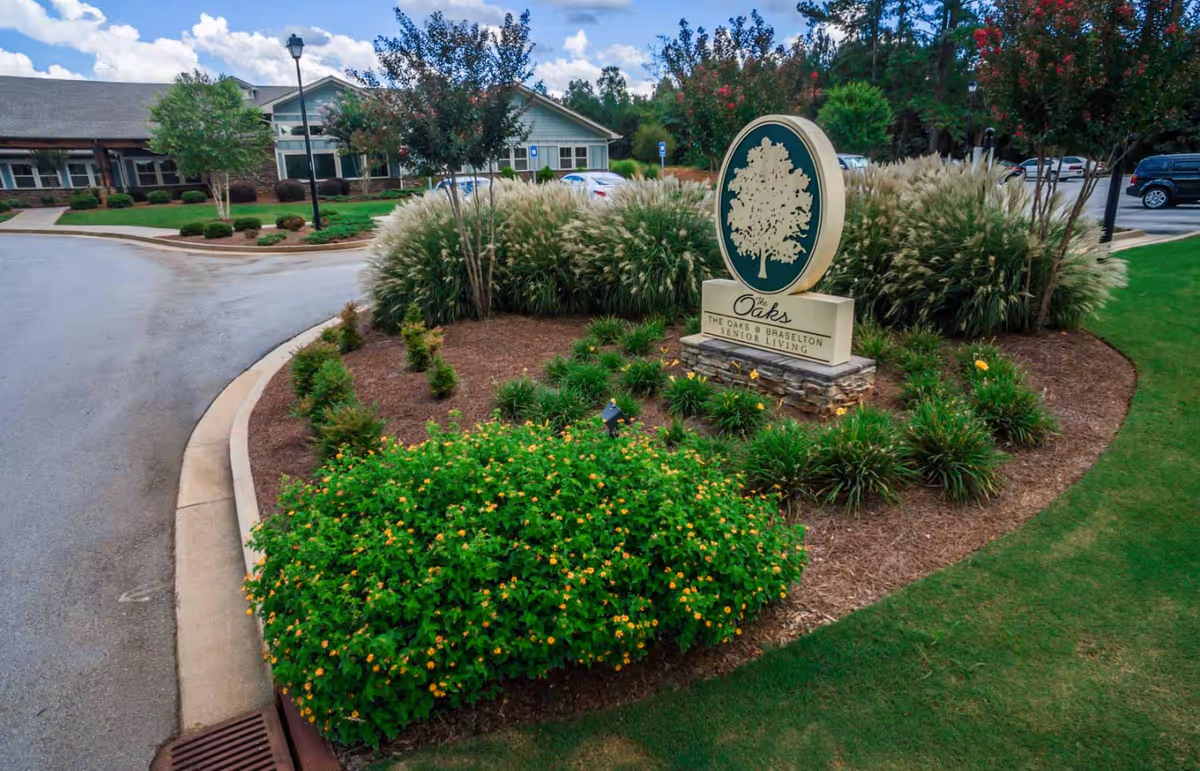 Landscaped roundabout with a sign reading 'The Oaks at Braselton Senior Living' in front of the facility building.