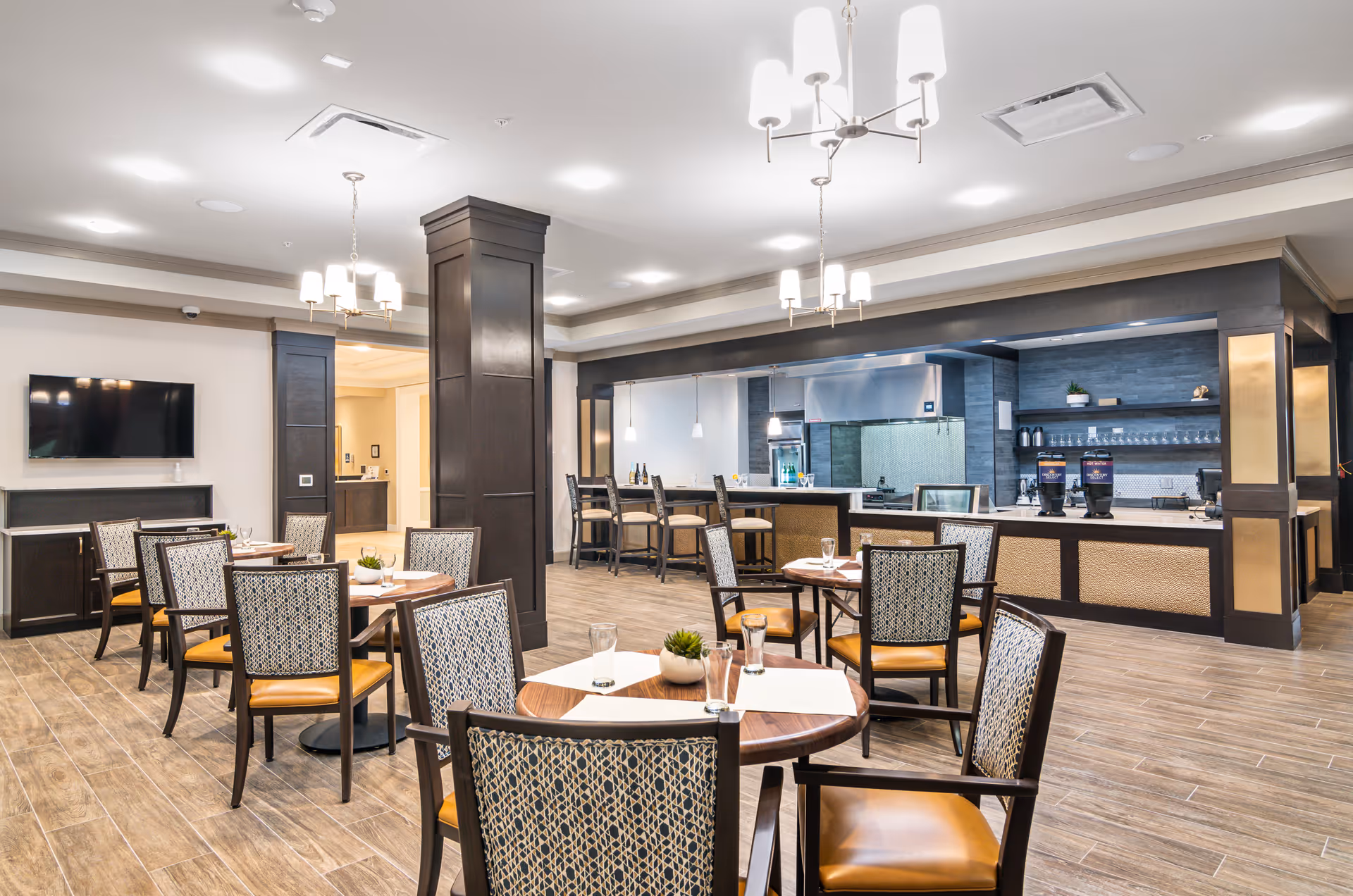 A modern dining area in a senior living facility featuring several round wooden tables with patterned chairs. The room has wood-look tile flooring, a flat-screen TV mounted on the wall, and a kitchen area with a counter and bar stools in the background. The space is well-lit with ceiling lights and hanging chandeliers.