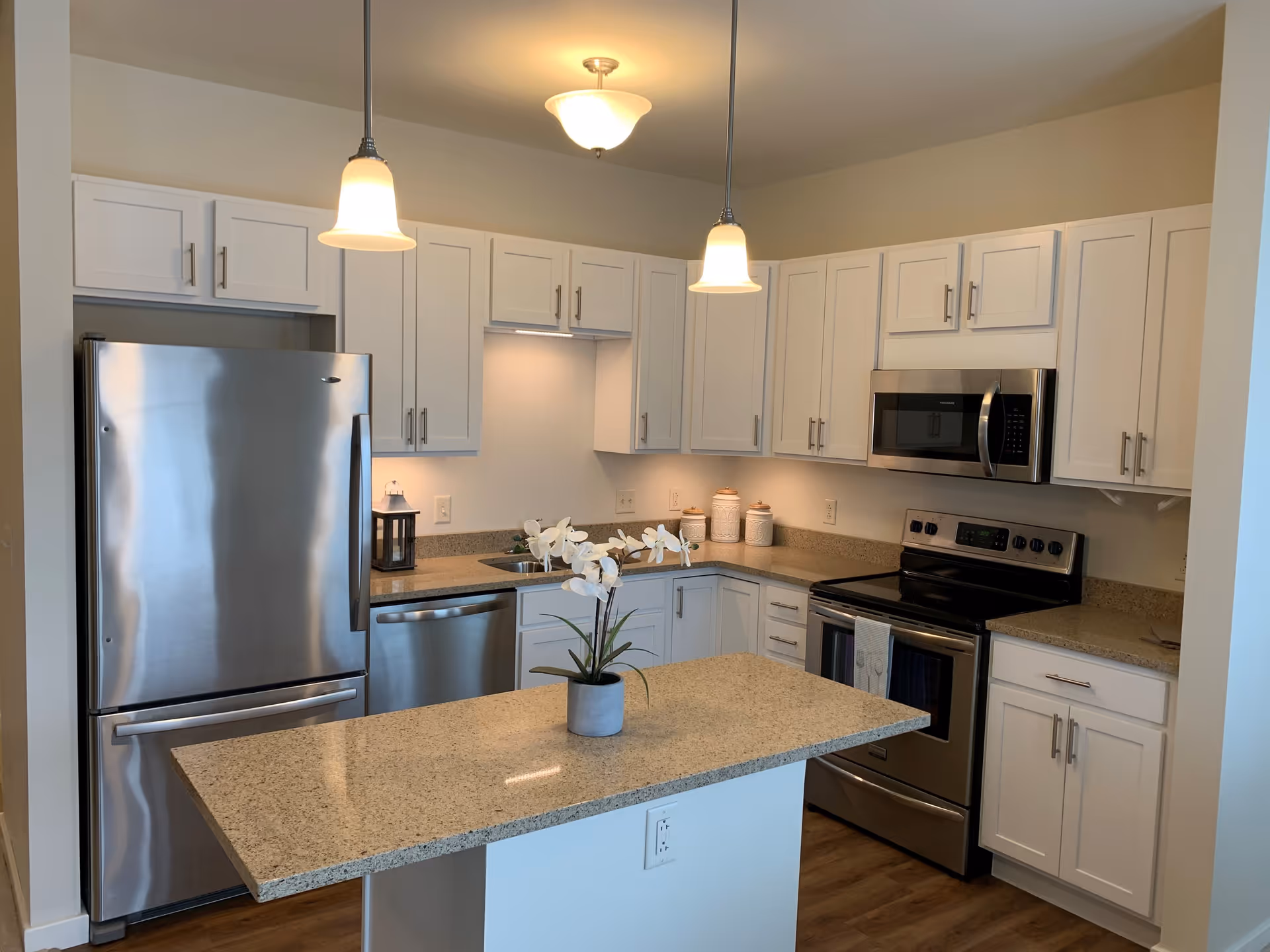 Modern kitchen with white cabinets, stainless steel refrigerator, dishwasher, oven, and microwave. There is a granite countertop island in the center with a small potted orchid plant. Two pendant lights hang above the island and a ceiling light is visible.