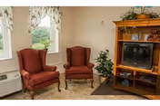 A cozy living room area with two red upholstered armchairs positioned near two windows with floral curtains. There is a wooden entertainment center holding an older-style television, some decorative items, and a potted plant next to it. The room has light-colored walls and carpeted flooring.