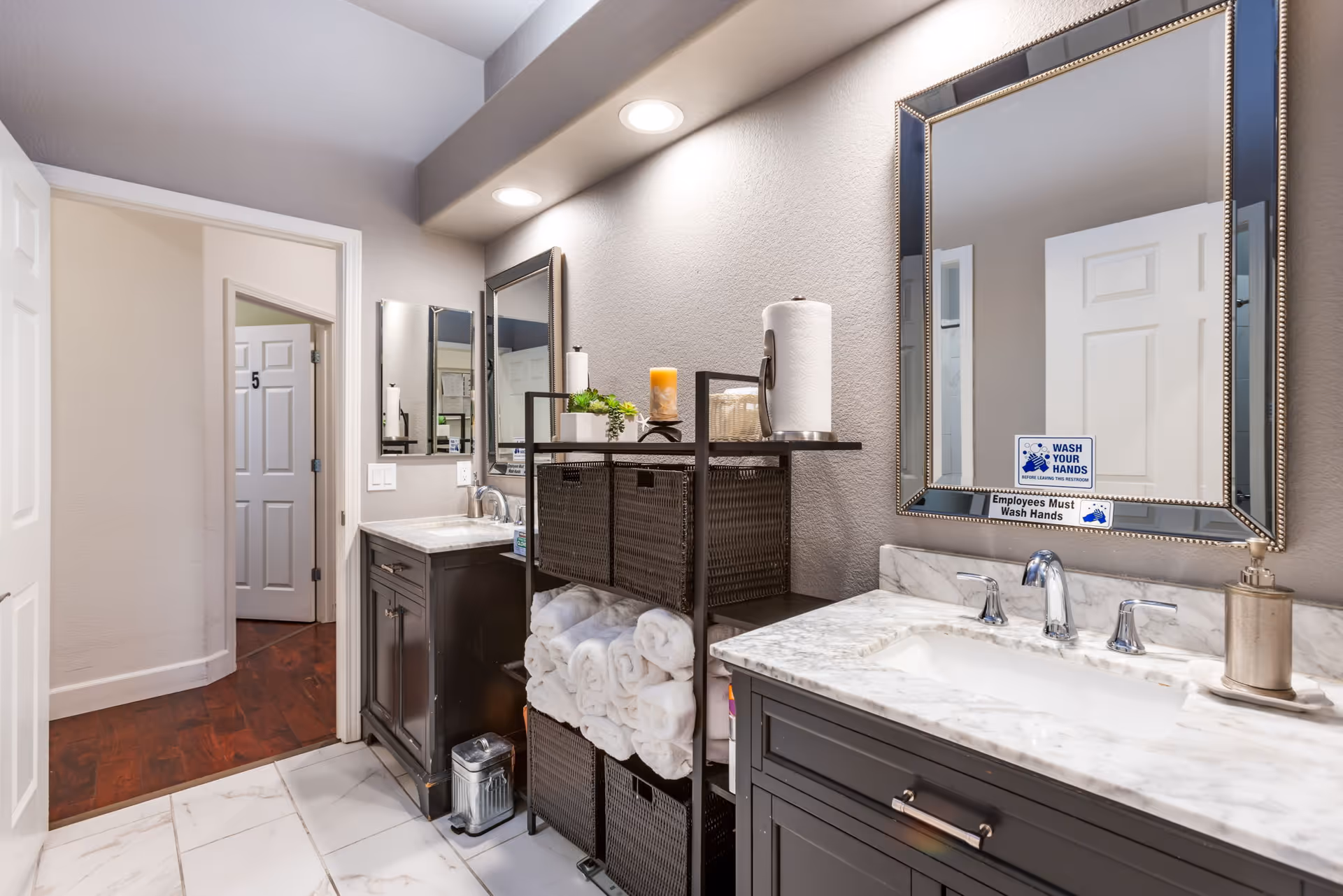 A clean and well-lit bathroom with two dark wood vanities topped with white marble countertops and silver faucets. Above each vanity is a large mirror, one of which has a sign that reads 'Employees Must Wash Hands.' Between the vanities is a black shelving unit holding rolled white towels, baskets, a paper towel roll, a candle, and a small plant. The floor is tiled in white, and an open door reveals a hallway with wooden flooring and a door labeled with the number 5.