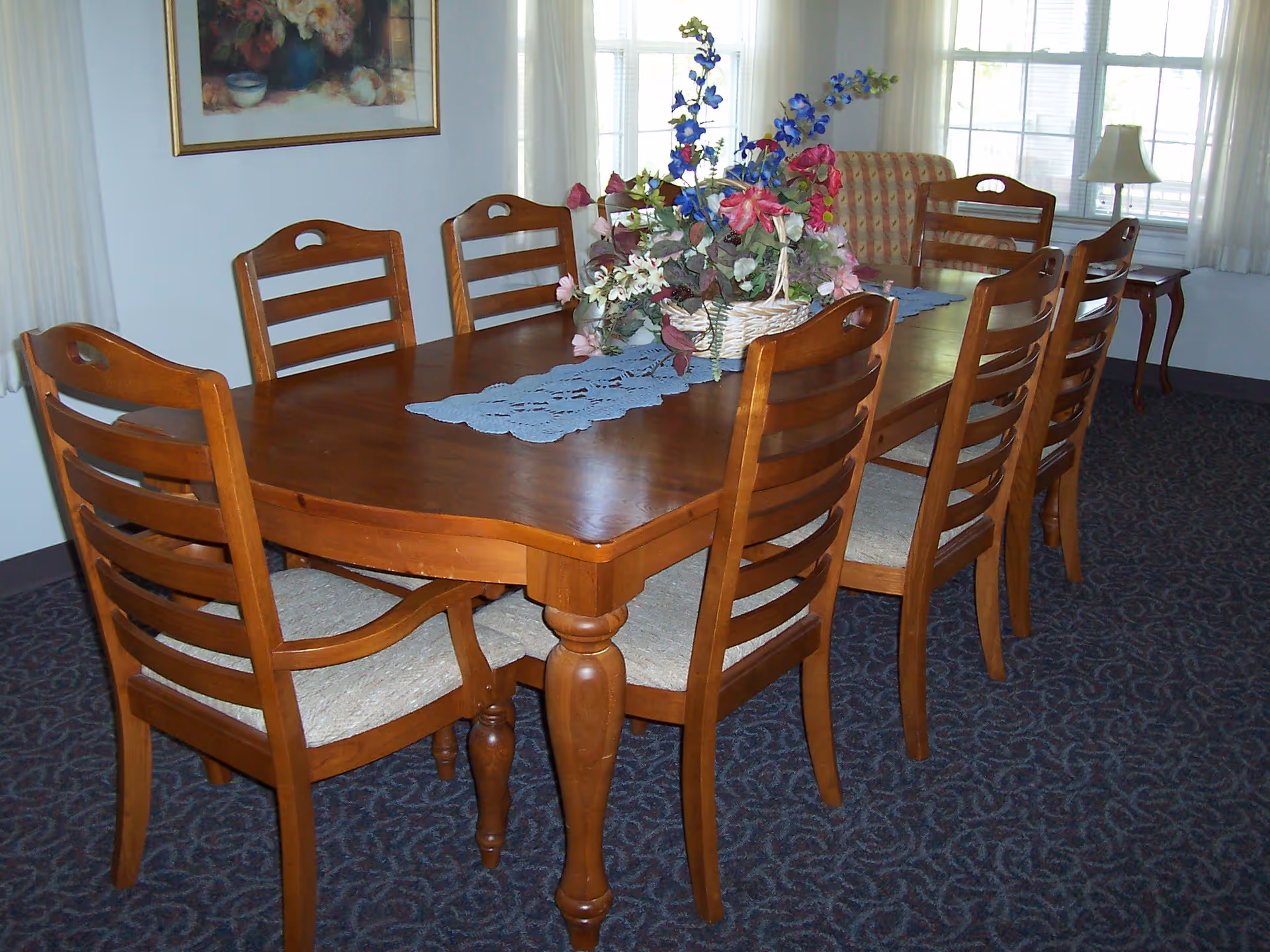A dining room with a wooden table surrounded by eight wooden chairs with cushioned seats. The table has a blue lace runner and a large floral centerpiece. In the background, there is a window with white curtains, a framed floral painting on the wall, and a cushioned armchair.