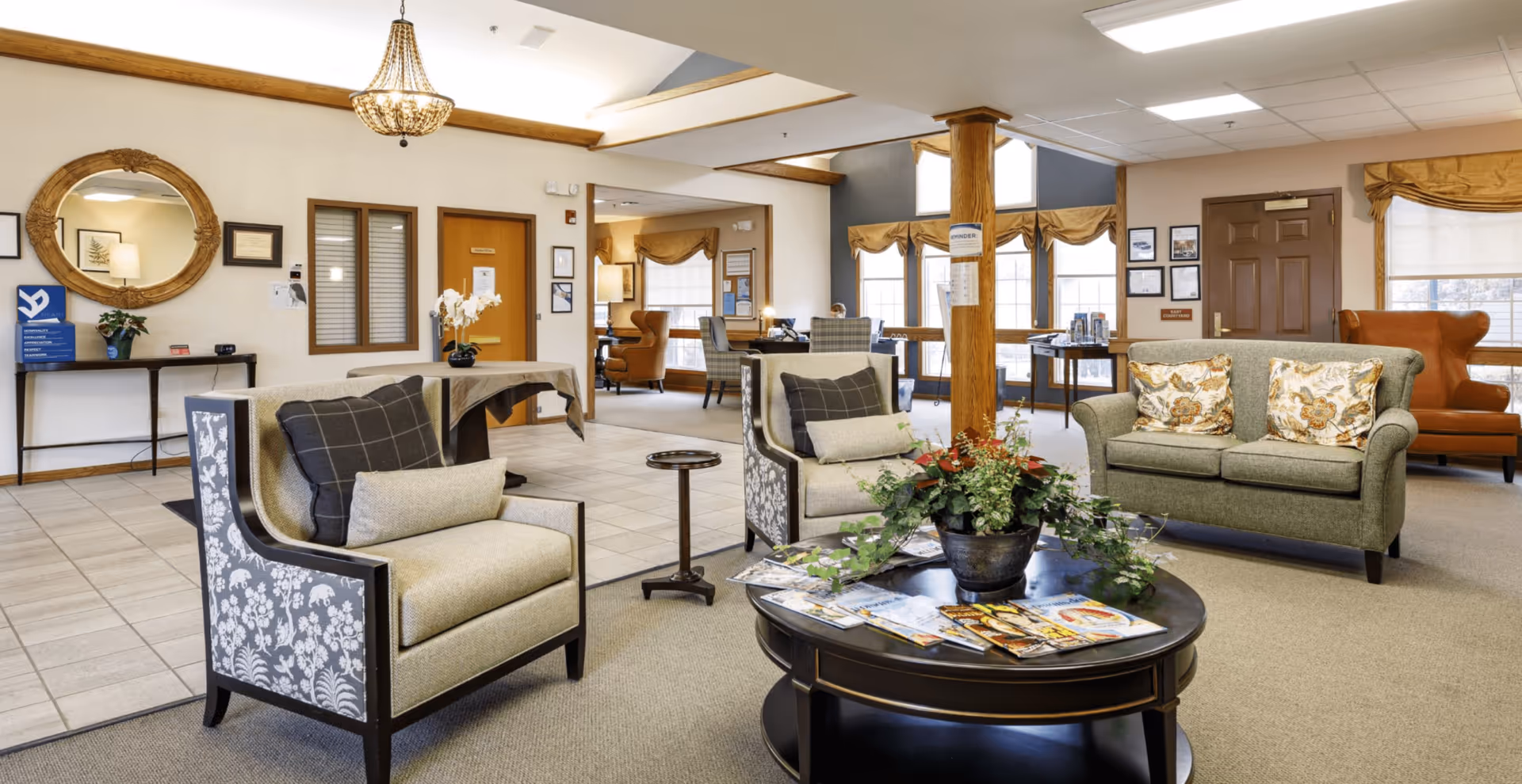 Bright, well-furnished senior living facility common area with armchairs and sofas arranged around a central coffee table topped with magazines and a plant, with reception and seating visible in the background.