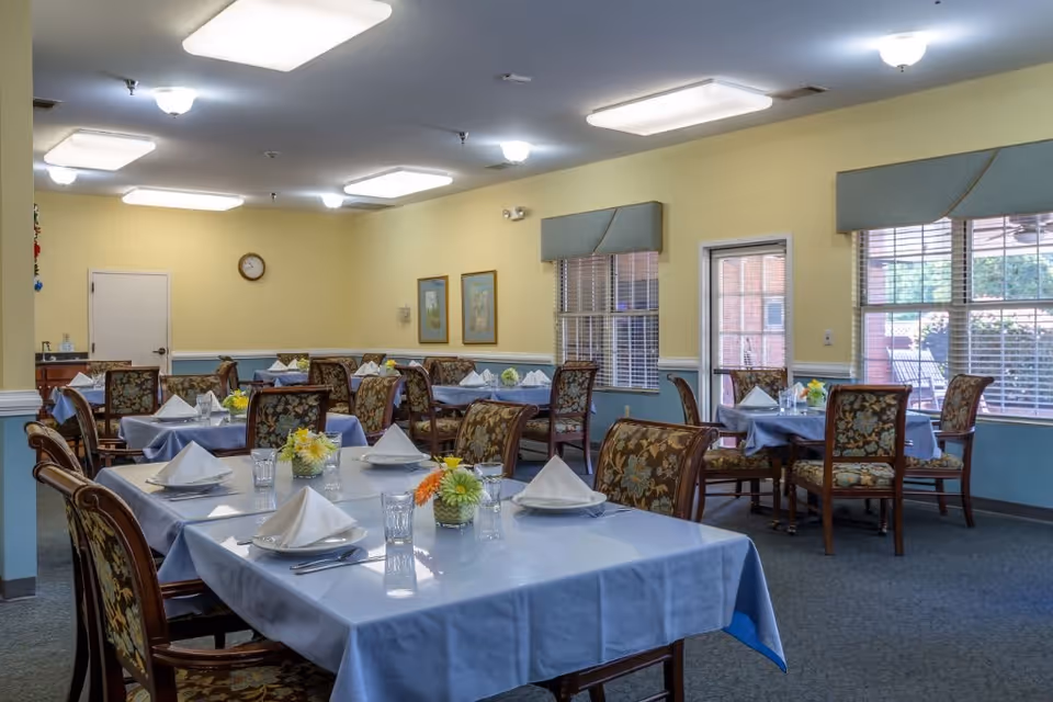 A dining room with several tables covered in blue tablecloths, each set with folded white napkins, glasses, and small flower arrangements. The room has yellow walls with blue wainscoting, floral upholstered chairs, large windows with blinds, and ceiling lights.
