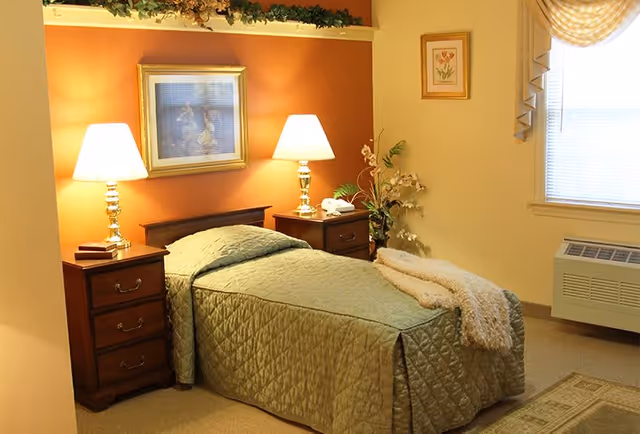 A cozy bedroom in Franciscan Manor featuring a single bed with a green quilted bedspread and a beige throw blanket. The bed is flanked by two wooden nightstands, each with a lamp. Above the bed hangs a framed picture, and there is a window with blinds and a valance on the right side. A small plant and a framed floral picture decorate the room.