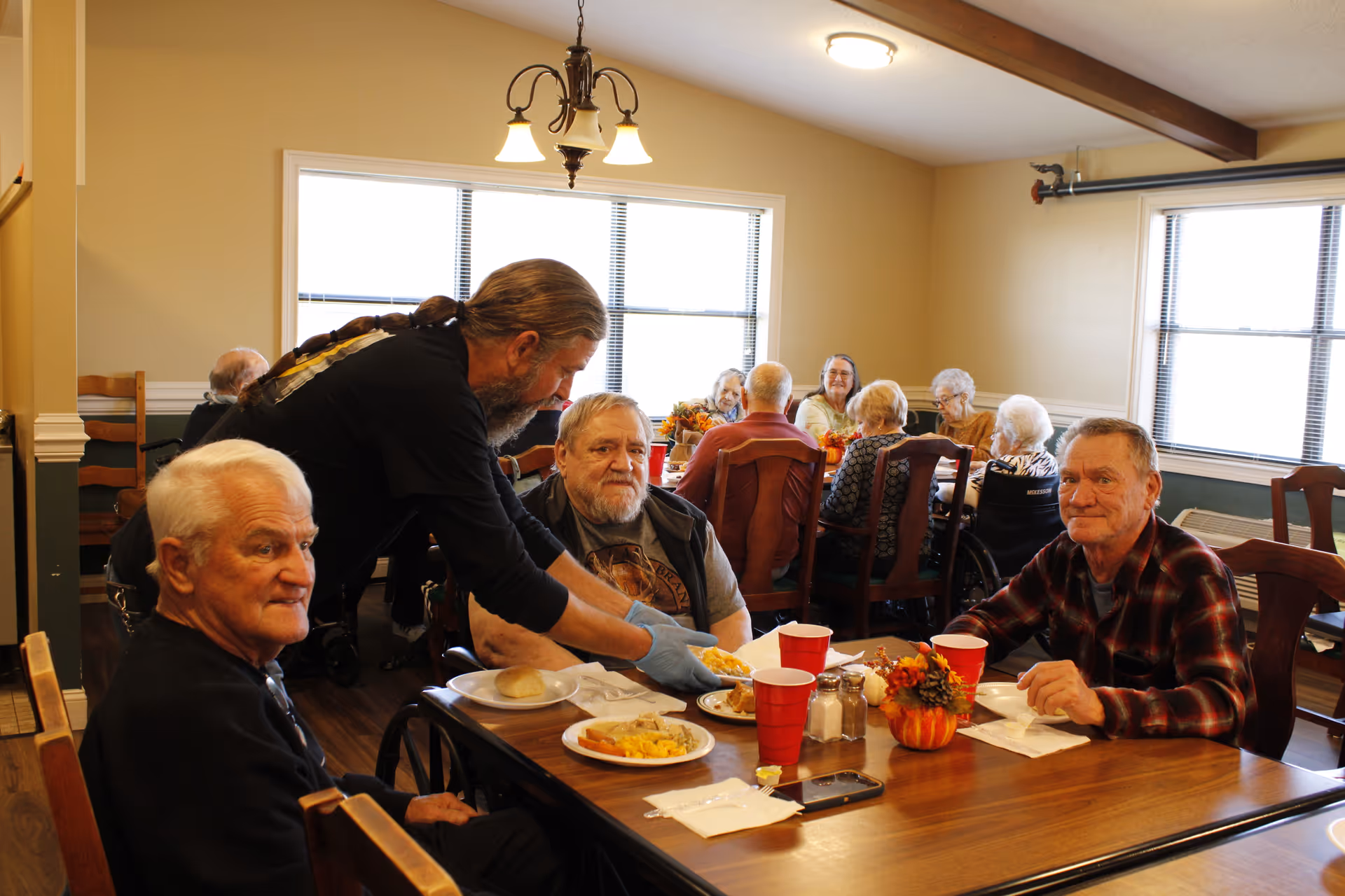 A group of elderly people sitting around tables in a dining room at Golden Oaks Assisted Living. A staff member wearing gloves is serving food to one of the men. The room has large windows, wooden chairs, and fall-themed decorations on the tables.