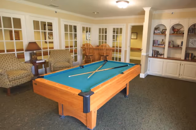 A recreational room with a pool table in the center, two crossed pool cues on the table, two patterned armchairs with a small side table and lamp between them, a built-in white bookshelf with decorative items and books, and a set of glass-paneled double doors leading to another room.