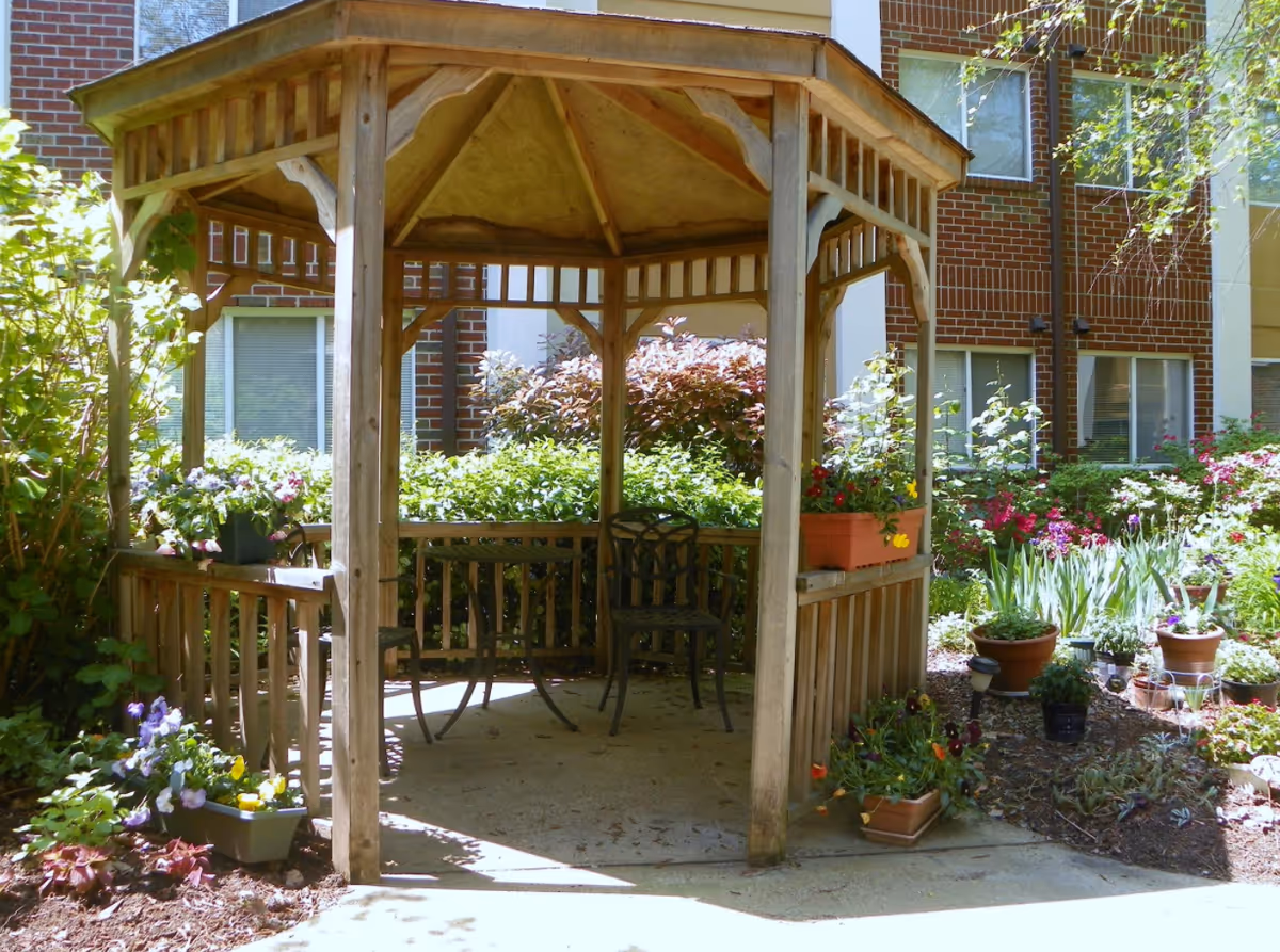 Wooden gazebo with chairs and flower planters in a landscaped courtyard in front of a brick building.