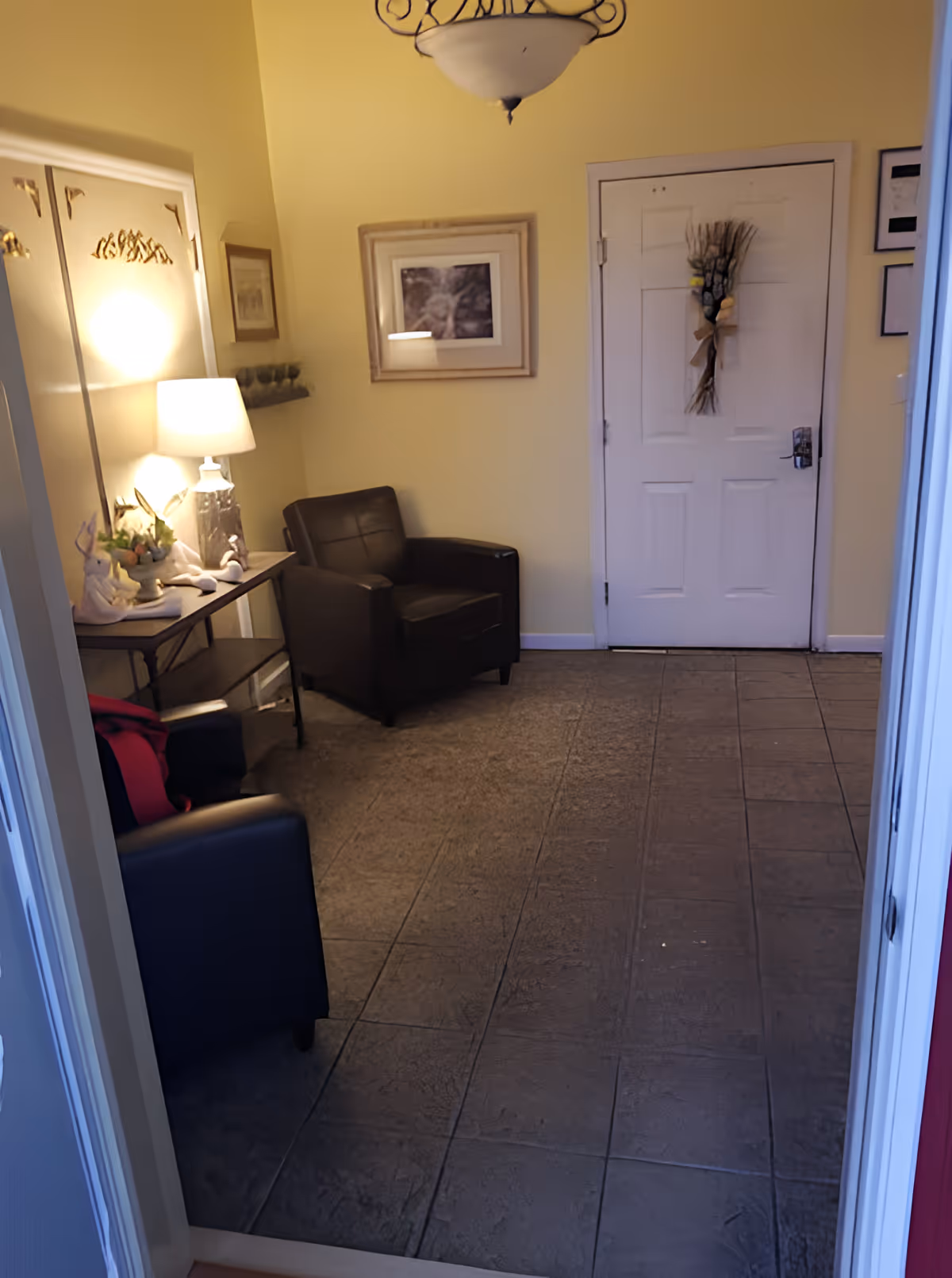 A small, cozy entryway with a white door decorated with a hanging floral arrangement. The walls are painted yellow, and there are two dark brown armchairs placed against the walls. A narrow table with a lamp, decorative items, and a small plant is positioned along the left wall. Above the table is a framed picture and a small shelf with glassware. The floor is tiled with a textured pattern, and a ceiling light fixture is visible.