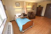 A senior care patient room with a single hospital-style bed with a blue blanket, a brown recliner, a wooden cabinet, and wood-look flooring.