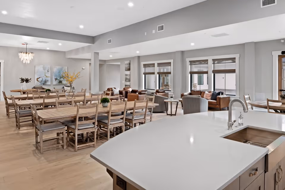 A spacious and modern communal dining and living area in Cadence Independent Living. The room features multiple wooden dining tables with chairs, a large kitchen island with a sink in the foreground, and a comfortable seating area with sofas and armchairs near windows with blinds. The space is well-lit with recessed lighting and decorated with vases of flowers and abstract wall art.