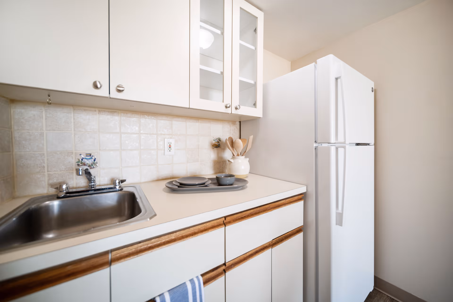 A small kitchen area with a white countertop, a stainless steel sink, white cabinets with wooden handles, a white refrigerator, and a few kitchen utensils in a white container on the counter. The backsplash is tiled in a light beige color.