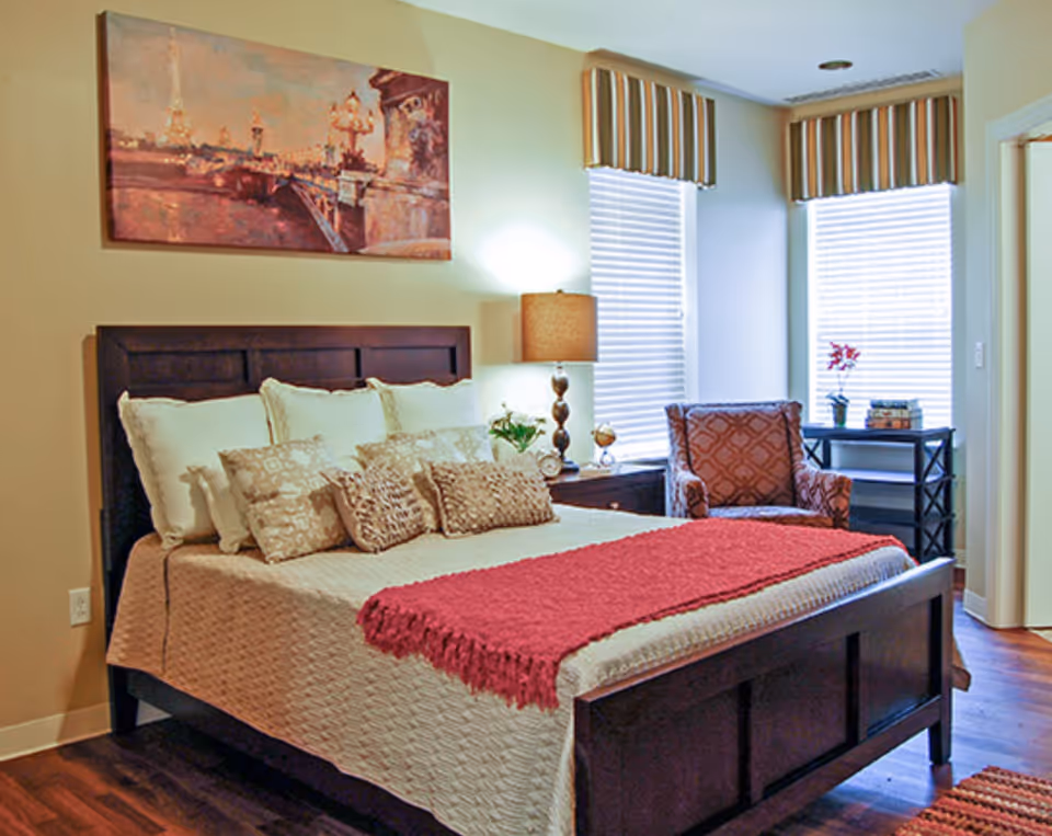 A cozy bedroom with a dark wooden bed frame, a beige quilted bedspread, multiple decorative pillows, and a red throw blanket. There is a bedside table with a lamp and a small plant. Two windows with striped valances let in natural light. An upholstered armchair and a small black shelving unit with books and a flower vase are near the windows. A painting depicting a cityscape hangs above the bed.