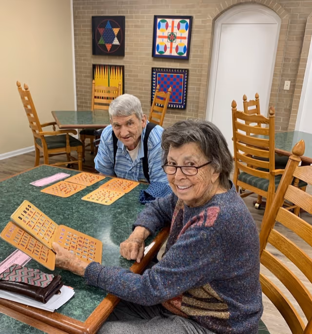 Two elderly individuals sitting at a green table playing bingo in a room with wooden chairs and colorful geometric artwork on a brick wall.