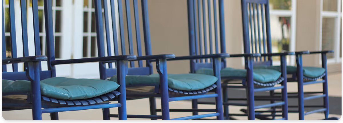 Row of blue wooden rocking chairs with teal cushions on a covered porch in front of a building.