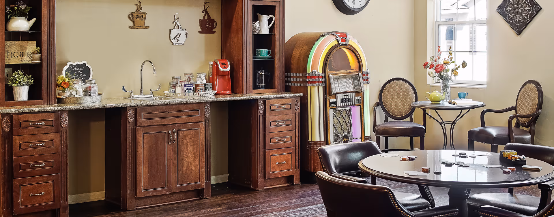 A cozy common area with a wooden kitchenette featuring a sink, coffee maker, and jars of condiments. There is a vintage jukebox next to the kitchenette. In front, there is a round table with chairs around it, some playing cards and poker chips on the table. A smaller round table with two chairs is placed near a window with a vase of flowers and a teapot on it. The room has warm lighting and decorative wall art.