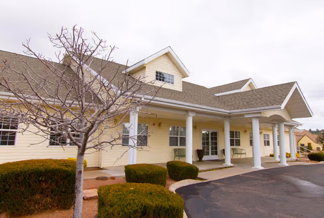 Exterior view of a single-story senior living facility building with a covered entrance supported by white columns. The building is light yellow with white trim and has a gabled roof. There are trimmed bushes and a leafless tree in the landscaped area near the entrance.