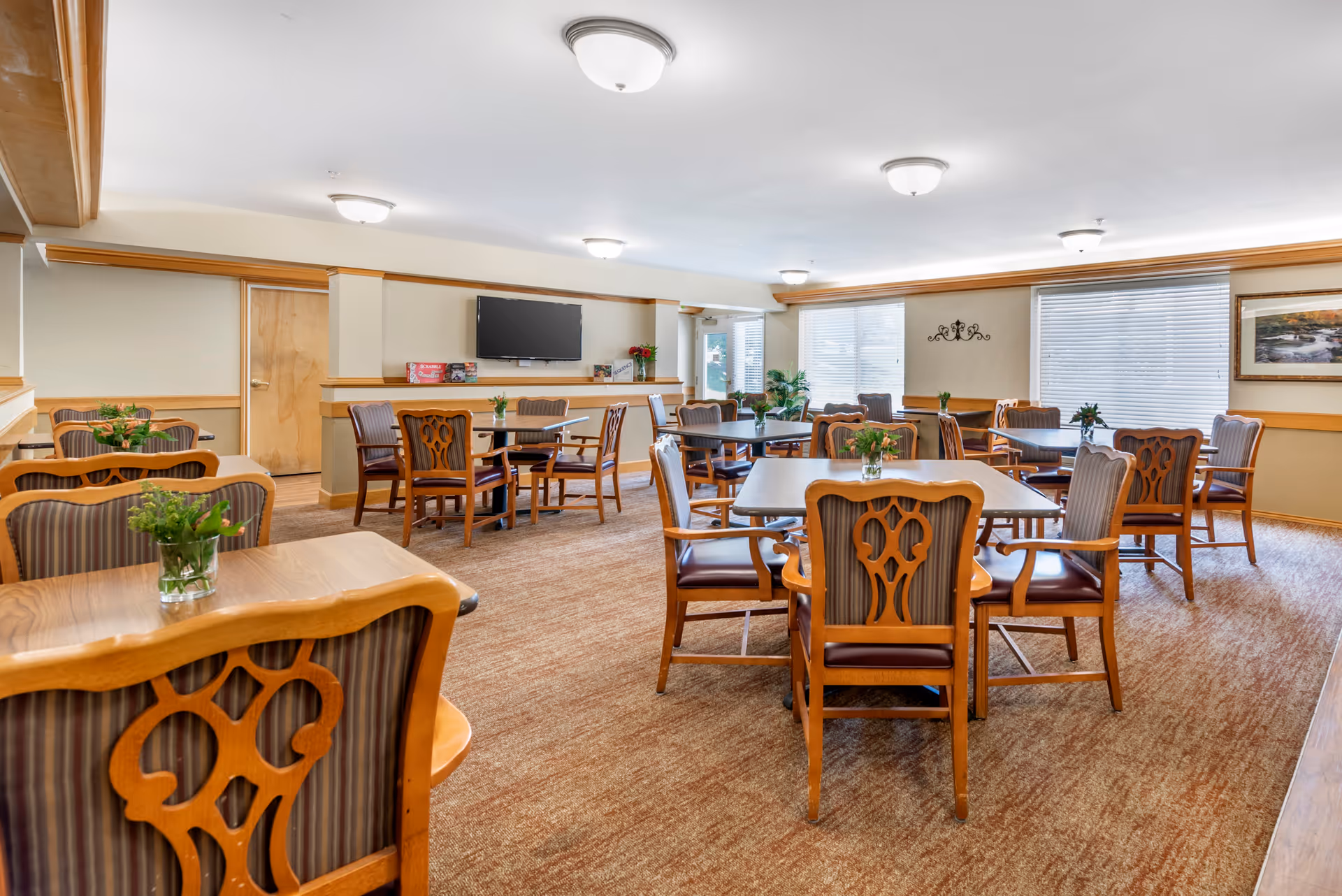 Bright communal dining room with wooden tables and chairs, small potted plants, and a wall-mounted TV.