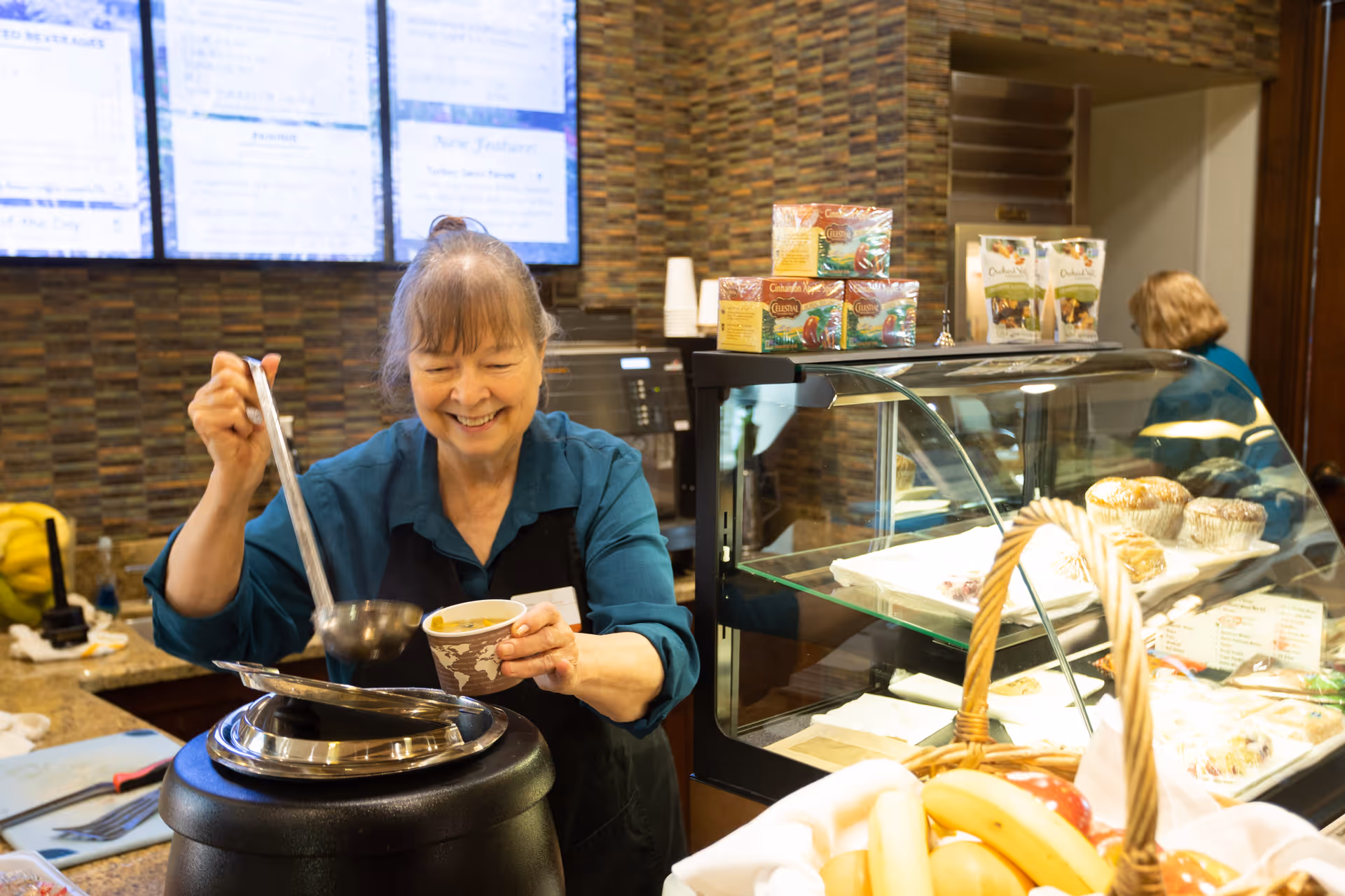 A smiling woman wearing a teal shirt and black apron ladles soup into a paper cup behind a counter in a food service area. In front of her is a soup warmer, and to the right is a glass display case with muffins and other baked goods. A basket of fruit is visible in the foreground, and a menu board is on the wall behind her.