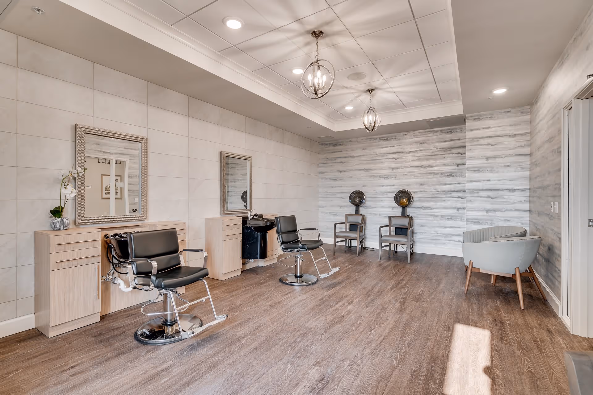 Interior view of a senior living facility hair salon with two black salon chairs in front of mirrors and sinks, two hair dryers with chairs against a textured wall, and two light gray armchairs in the corner on wood flooring.