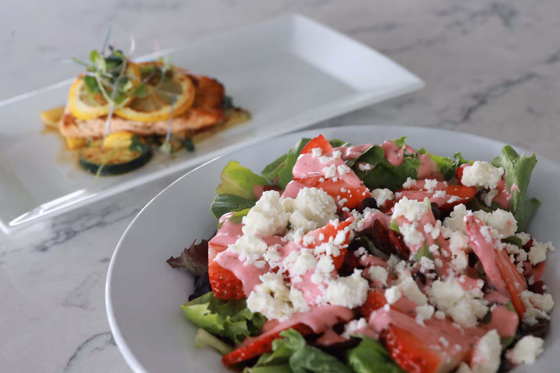 A close-up view of a fresh salad with leafy greens, strawberries, crumbled cheese, and pink dressing in a white bowl, with a plate of cooked salmon topped with lemon slices and microgreens in the background on a marble surface.