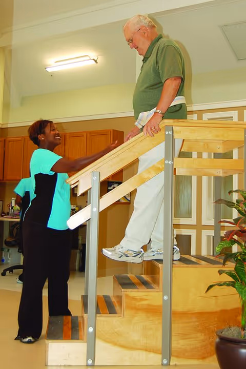 An elderly man wearing a green shirt and white pants is walking up a wooden staircase with handrails inside a facility. A smiling female caregiver in a turquoise and black uniform is standing beside him, offering support. The background shows wooden cabinets and a well-lit interior space.