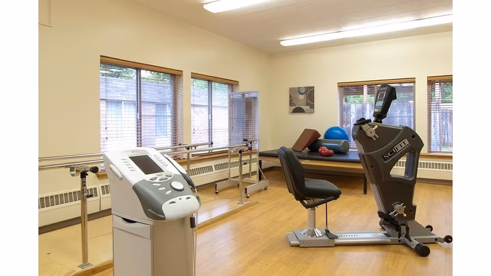 A physical therapy room with exercise equipment including a seated exercise machine, parallel bars for walking support, and a therapy table with exercise balls and a foam roller. The room has large windows with blinds and light-colored walls and flooring.
