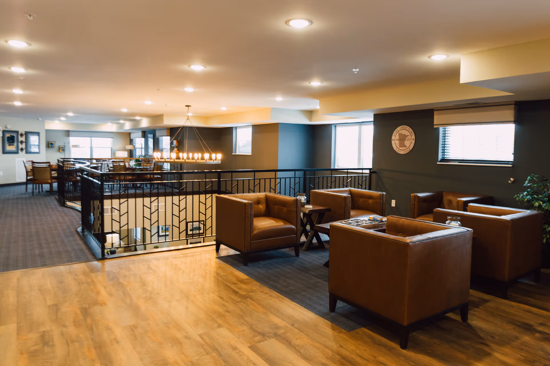 Interior view of a senior living facility lounge area with four brown leather armchairs arranged around a small wooden table. The space has wooden flooring, carpeted sections, modern lighting, and large windows allowing natural light. A decorative wall clock and some plants are visible on the walls.