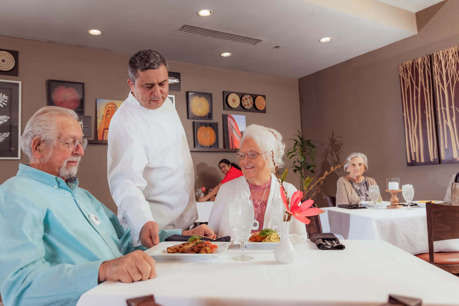 An elderly couple being served a meal by a waiter in a dining room with other residents at nearby tables.