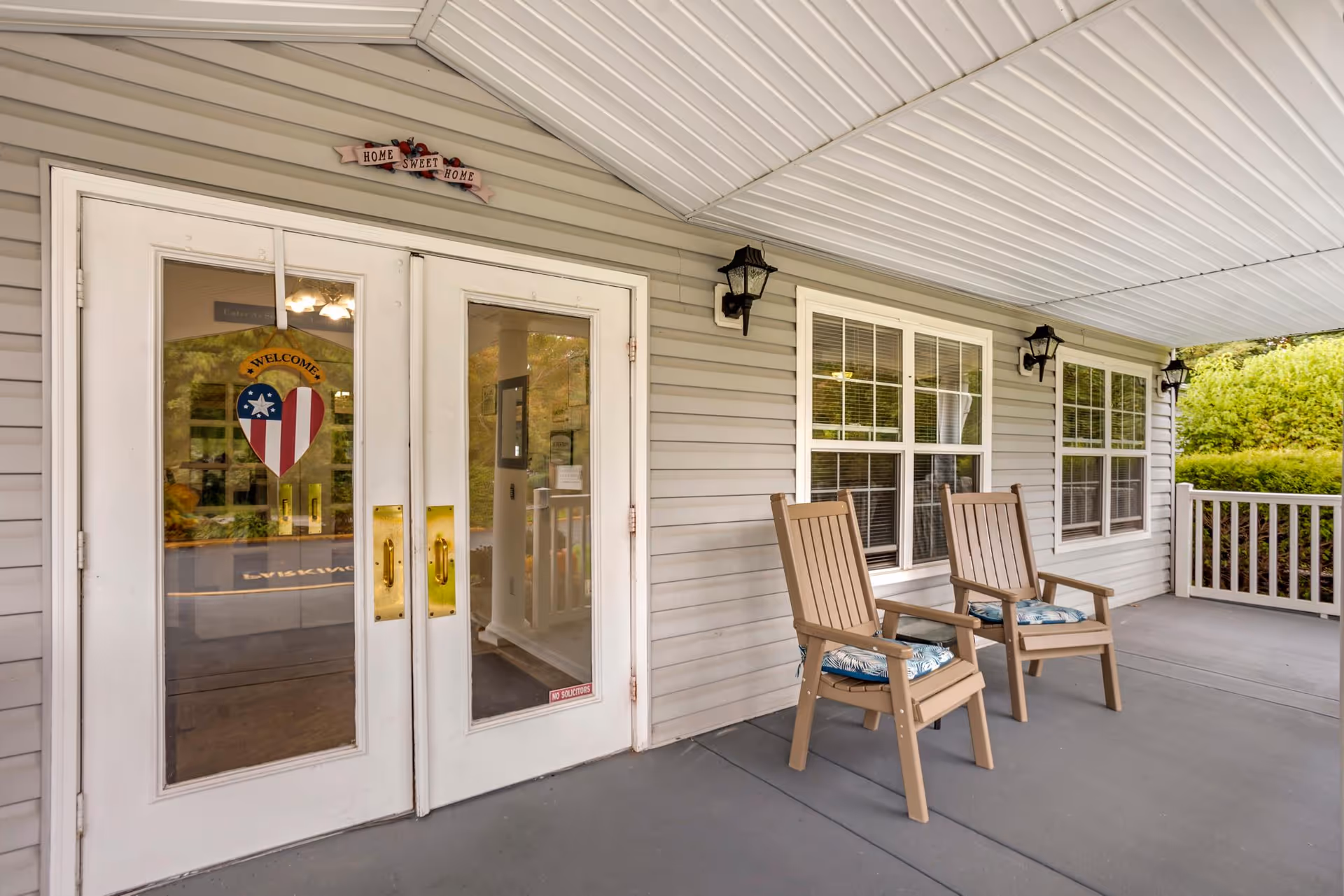 Covered porch area with two wooden chairs with cushions, white double doors with glass panels and a 'Welcome' heart-shaped decoration, beige siding, three black wall-mounted lantern lights, and a 'Home Sweet Home' sign above the doors.