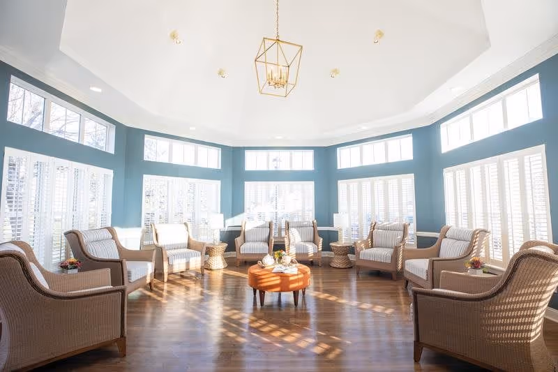 A bright, spacious sitting area with multiple wicker armchairs arranged in a circle around a round orange ottoman. The room features large windows with white shutters allowing natural light to flood in, teal blue walls, and a high white ceiling with a modern gold chandelier.