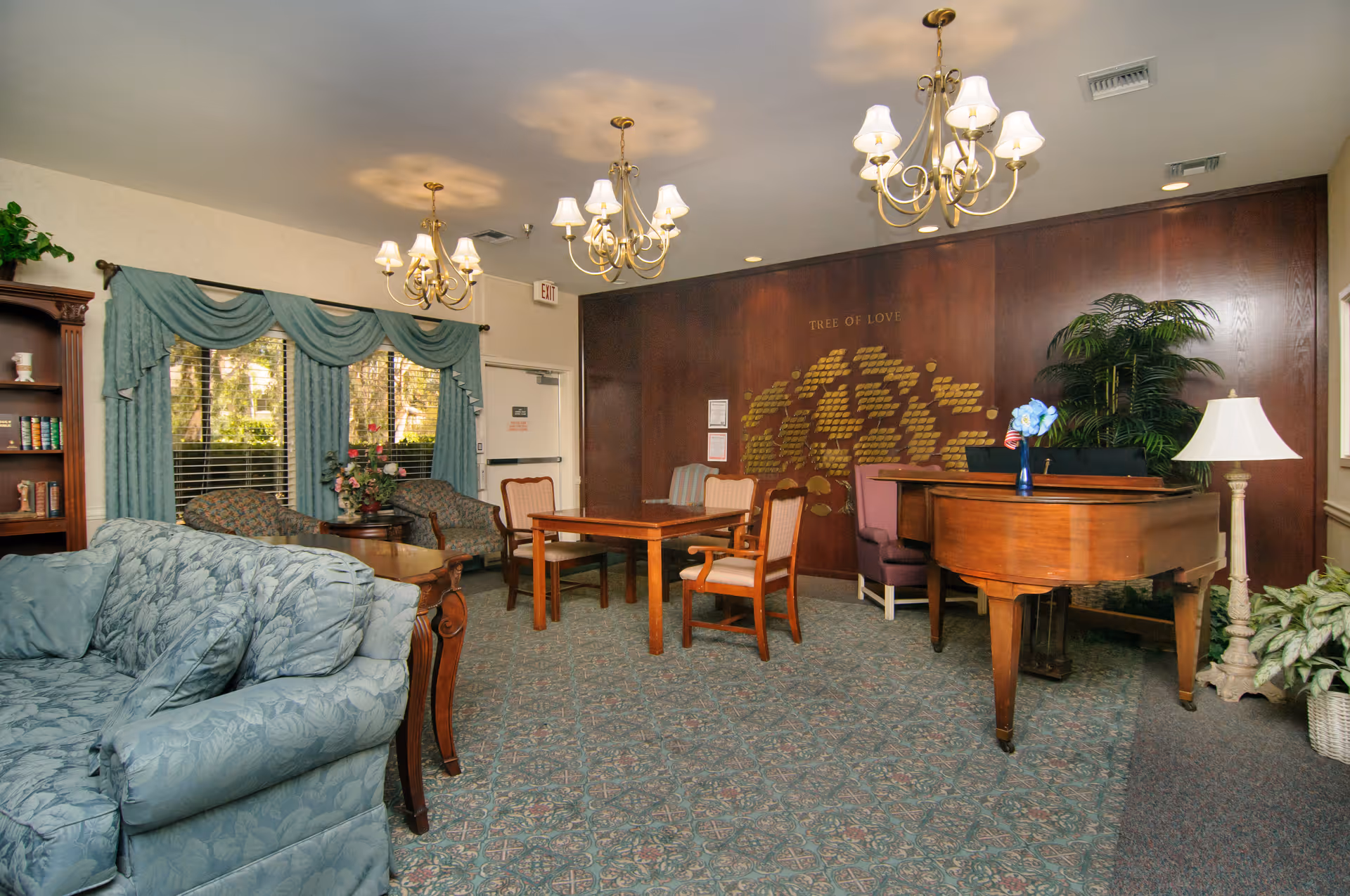 A cozy common area in a senior living facility featuring a blue patterned sofa, wooden chairs around a table, a grand piano with a vase of flowers, a bookshelf, and large windows with teal curtains. The back wall has a wooden panel with a 'Tree of Love' display and several chandeliers hanging from the ceiling.