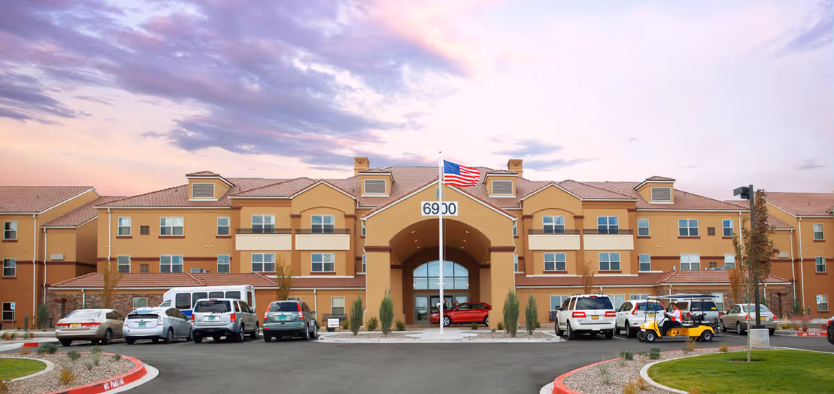 Three-story tan retirement building front with an arched main entrance, an American flag on a pole, and cars parked in front.