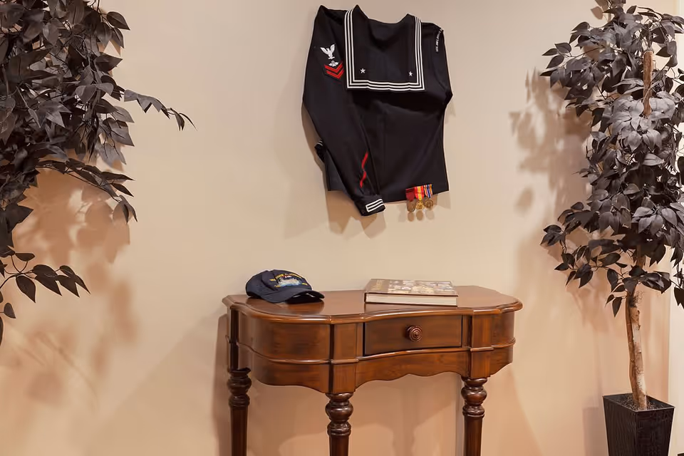 Wooden side table with a cap and book beneath a naval uniform top with medals on the wall, flanked by potted plants.