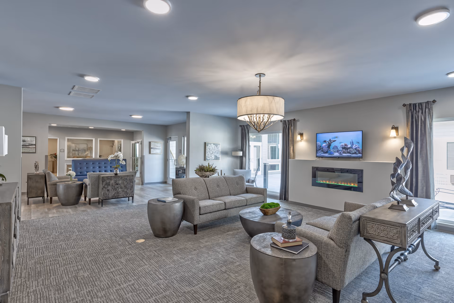 Spacious modern lounge with gray sofas, round side tables, a wall-mounted TV above an electric fireplace, and decorative lighting.