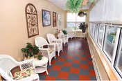 A bright sunroom with large windows on the right side, featuring white wicker chairs with floral cushions, small tables, potted plants, and decorative wall art on the left beige wall.