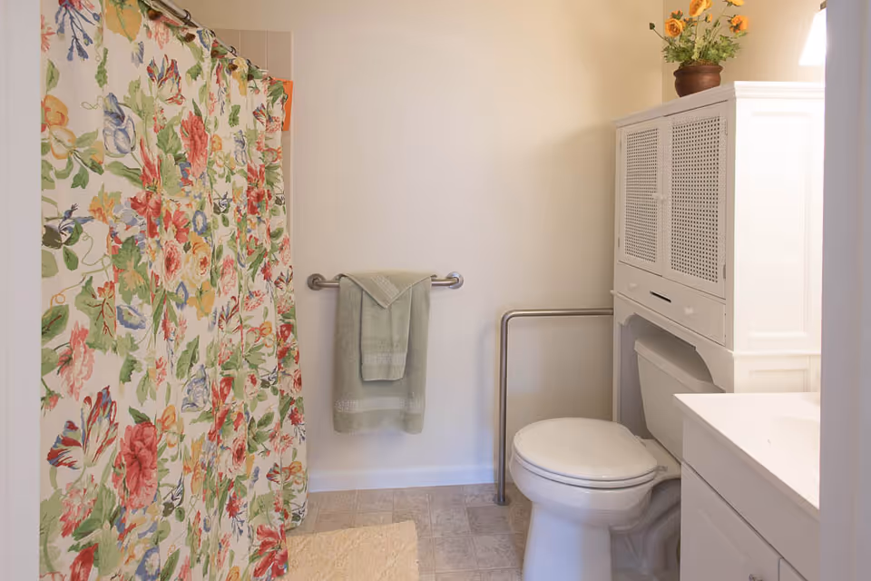 Bathroom featuring a floral shower curtain, toilet with safety grab bar, white storage cabinet, and a towel hanging on a bar.