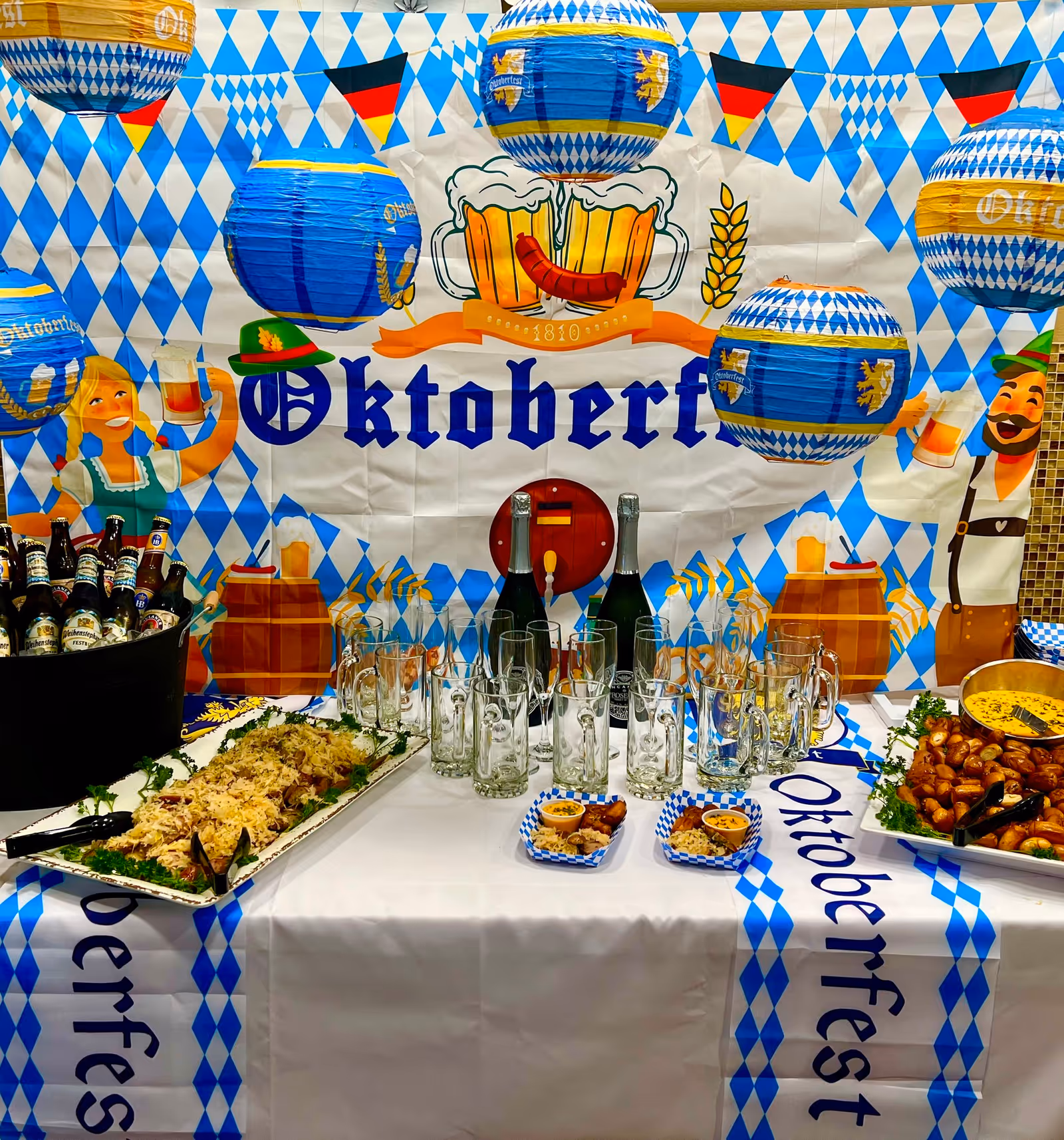 A festive Oktoberfest-themed table setup with blue and white diamond-patterned decorations, hanging lanterns, and a backdrop featuring cartoon characters holding beer mugs. The table is covered with a matching tablecloth and displays various food trays, beer bottles in a bucket, empty beer mugs, and small plates with snacks.