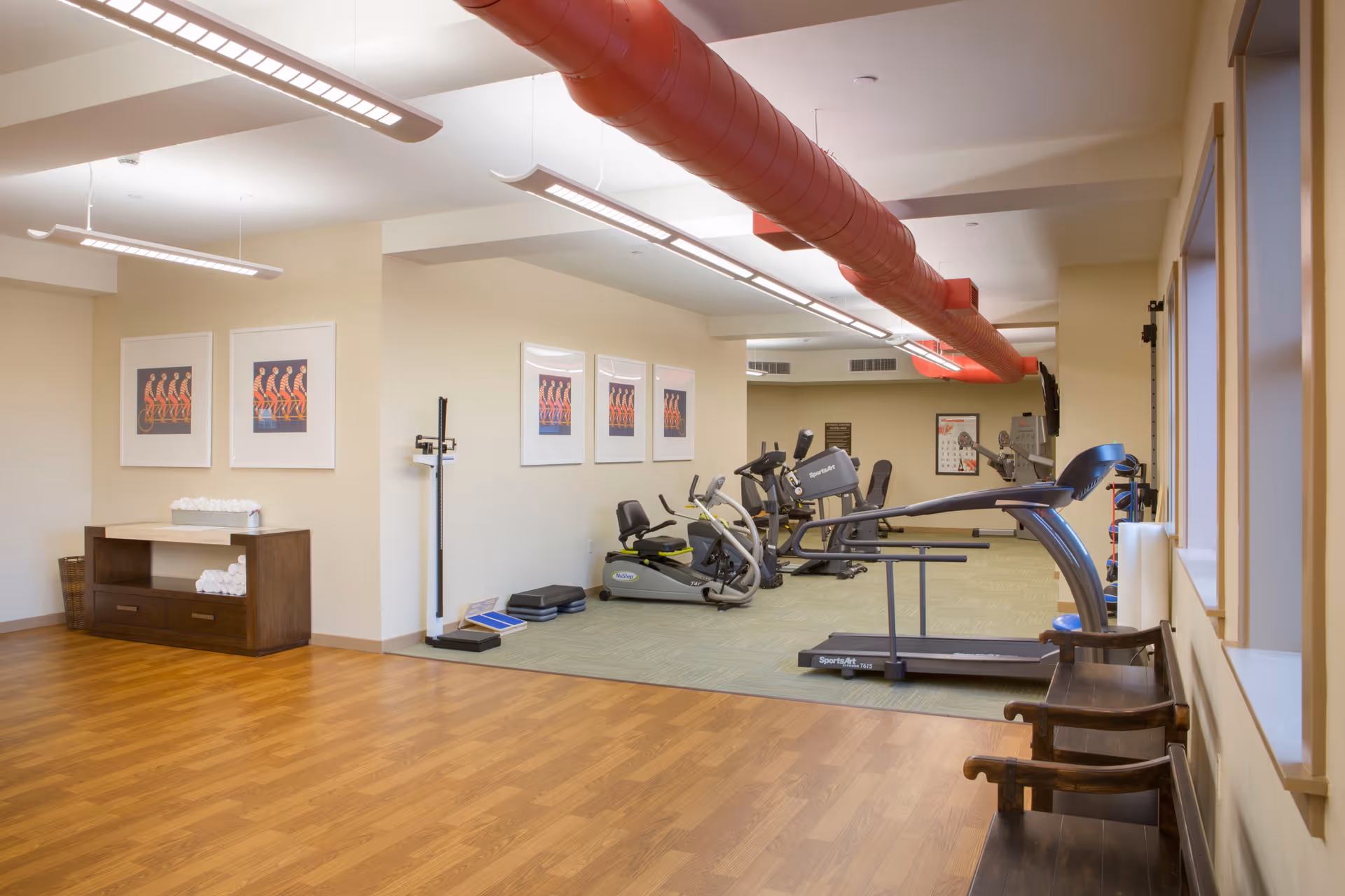 A fitness room in Prairie City Landing featuring exercise equipment including treadmills, stationary bikes, and elliptical machines. The room has wooden flooring in the foreground and carpeted area where the equipment is placed. There are framed pictures on the walls, a red exposed ventilation duct on the ceiling, and a wooden bench along the windowed wall.