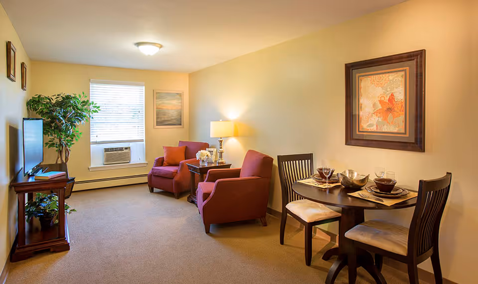 A cozy living room area with two red armchairs, a small wooden side table with a lamp and decorative items, a flat-screen TV on a wooden stand, a potted plant, and a window with blinds and an air conditioning unit. Adjacent to the seating area is a small round dining table set for two with dishes, glasses, and a decorative bowl. The walls are painted light yellow and decorated with framed artwork.