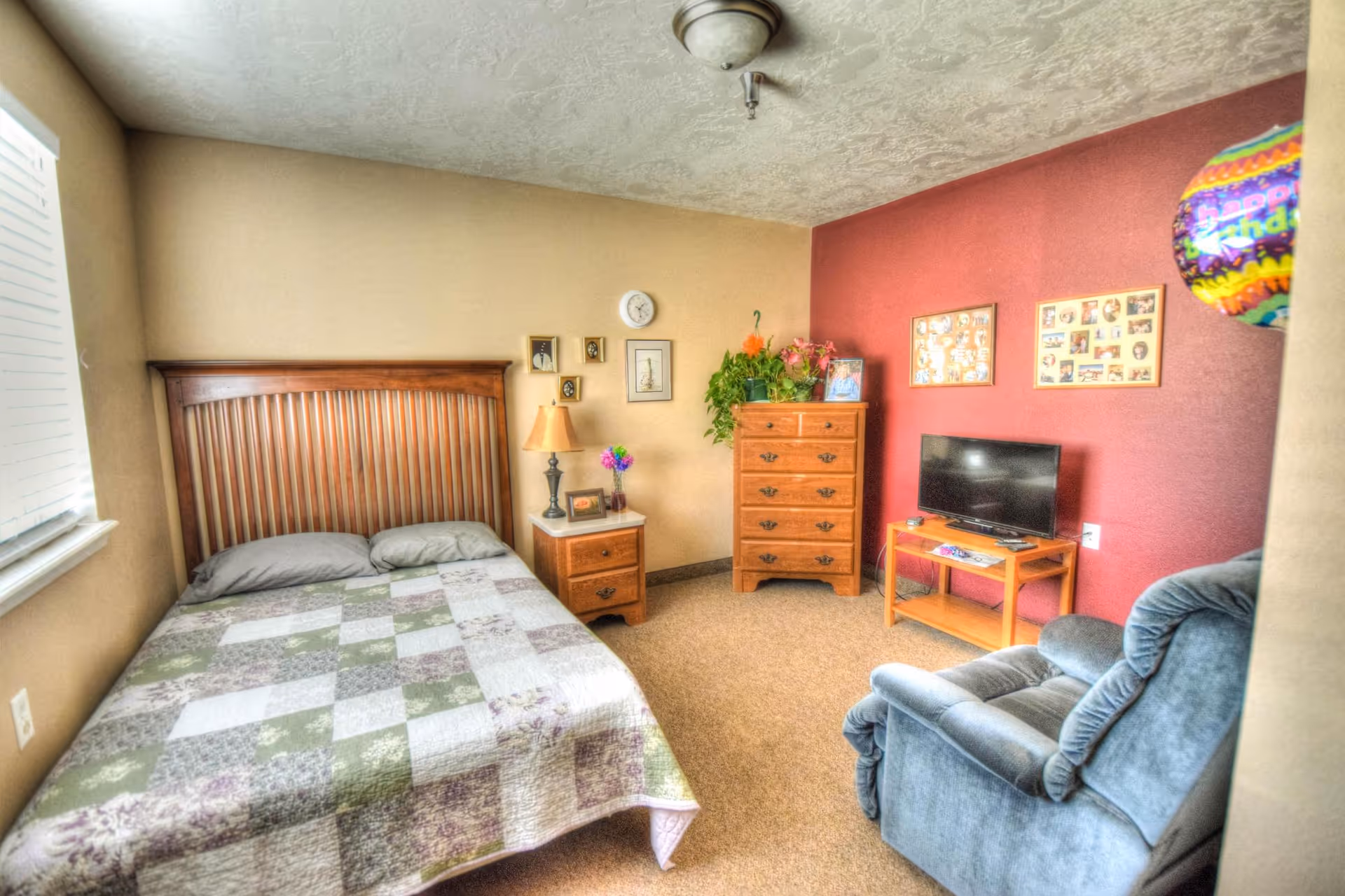 A cozy bedroom with a wooden bed frame and a patchwork quilt on the bed. There is a wooden nightstand with a lamp and a small vase of flowers next to the bed. On the wall above the nightstand are several framed pictures and a clock. Across the room is a wooden dresser with plants and framed photos on top. A flat-screen TV sits on a small wooden table against a red accent wall. A comfortable blue recliner chair is positioned near the TV. A colorful balloon is partially visible on the right side of the image.