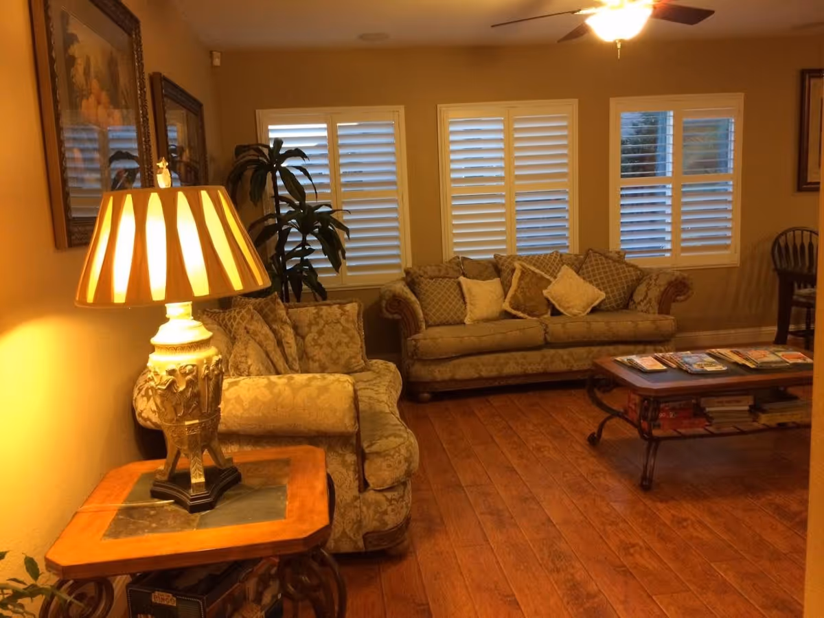 A cozy living room with two patterned sofas adorned with multiple cushions, a wooden coffee table with magazines on top, a side table with an ornate lamp, wooden flooring, and windows with white plantation shutters. There is a ceiling fan with a light fixture and framed artwork on the walls.