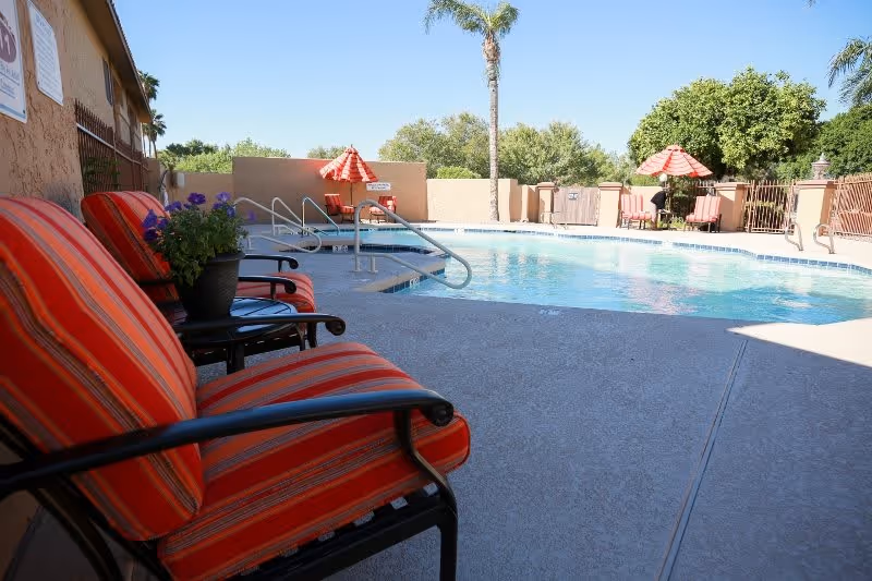 Outdoor swimming pool area with clear blue water, surrounded by a concrete deck. There are several red and orange striped cushioned chairs and matching umbrellas around the pool. Palm trees and green bushes are visible in the background under a clear blue sky.