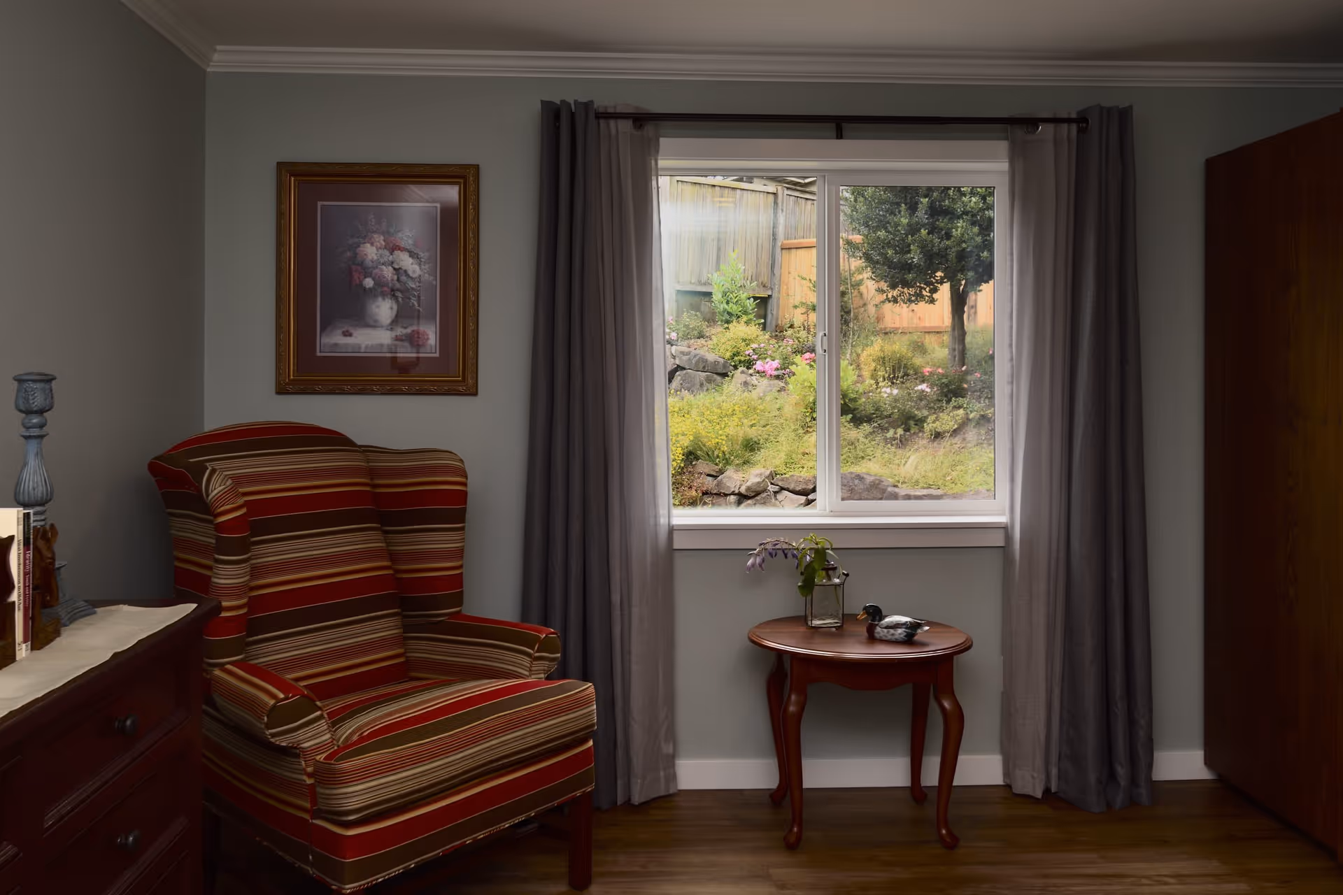 A cozy interior corner of a room featuring a striped upholstered armchair in red, beige, and brown tones next to a small wooden side table with a vase of flowers and a decorative duck figurine. A window with gray curtains reveals a garden with greenery, flowers, and a wooden fence outside. A framed floral painting hangs on the wall above the armchair, and a wooden dresser with books and a candle holder is partially visible on the left.