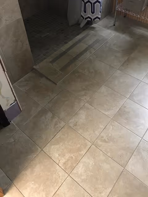 Tiled floor area with beige and light brown tiles leading to a tiled shower area with a glass door. A patterned blue and white towel hangs near the shower, and a metal rack is visible on the right side.
