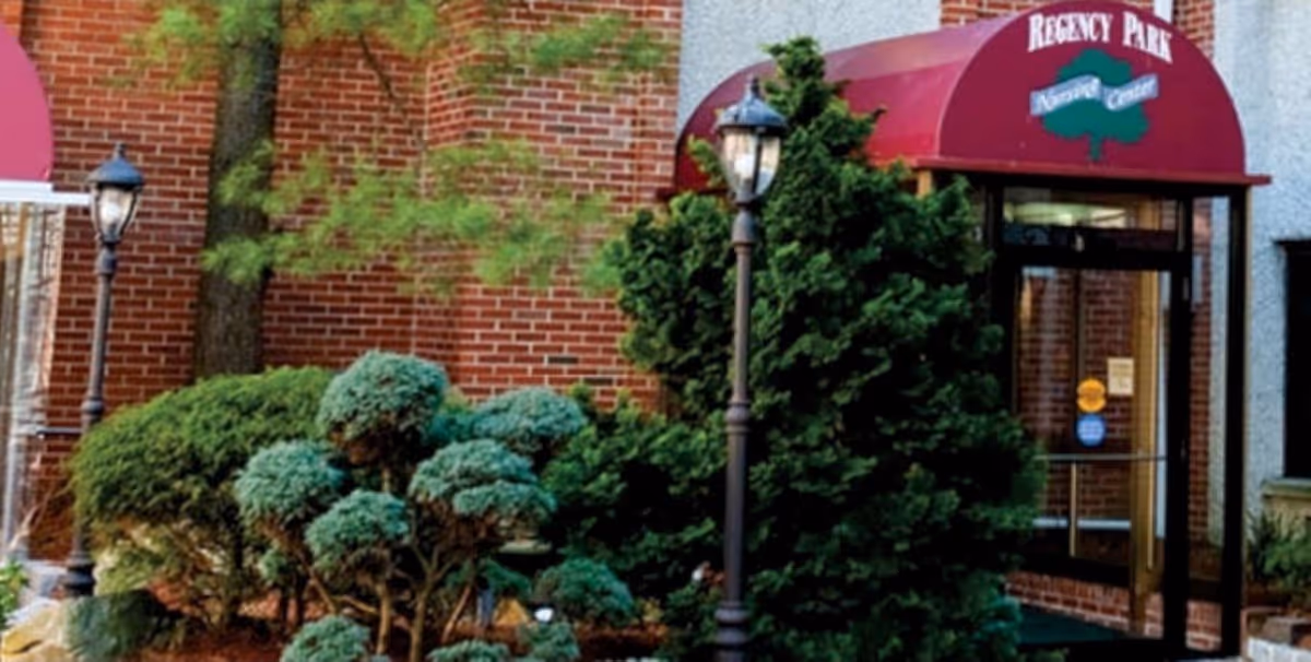 Entrance to a building with a red awning labeled 'Regency Park Recovery Center,' surrounded by green bushes and trees, with two black lamp posts in front.