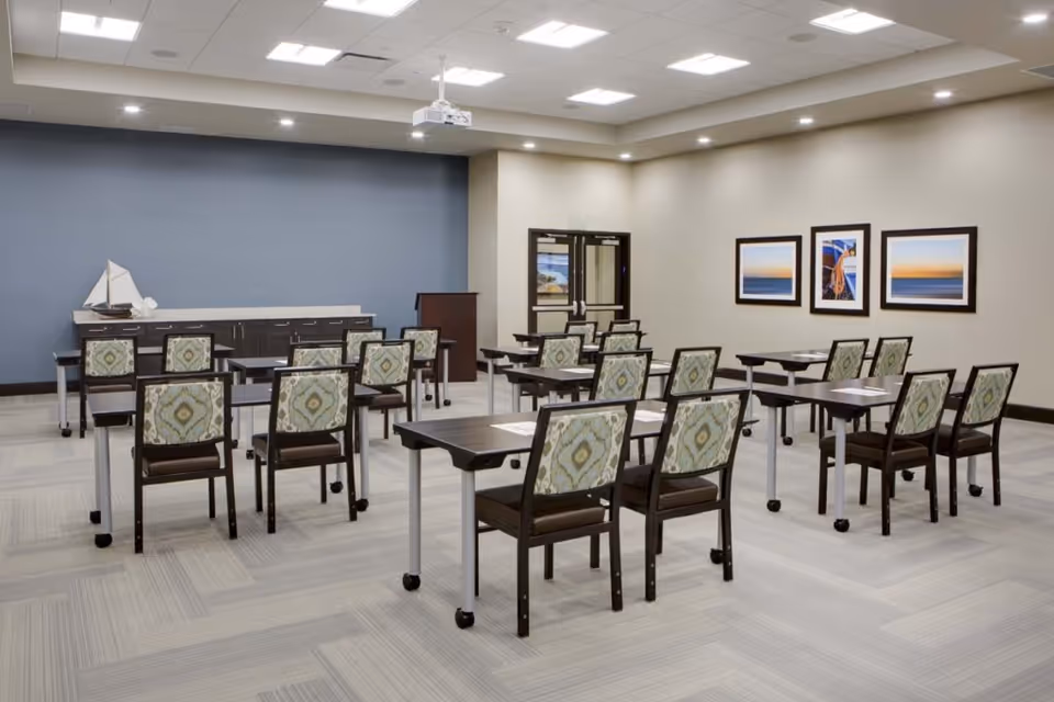 A well-lit room with multiple tables and chairs arranged in rows facing a podium and a cabinet against a blue accent wall. The chairs have patterned upholstery, and there are three framed pictures on the beige wall to the right. The room has a light-colored carpet and a ceiling with recessed lighting and a projector mounted.